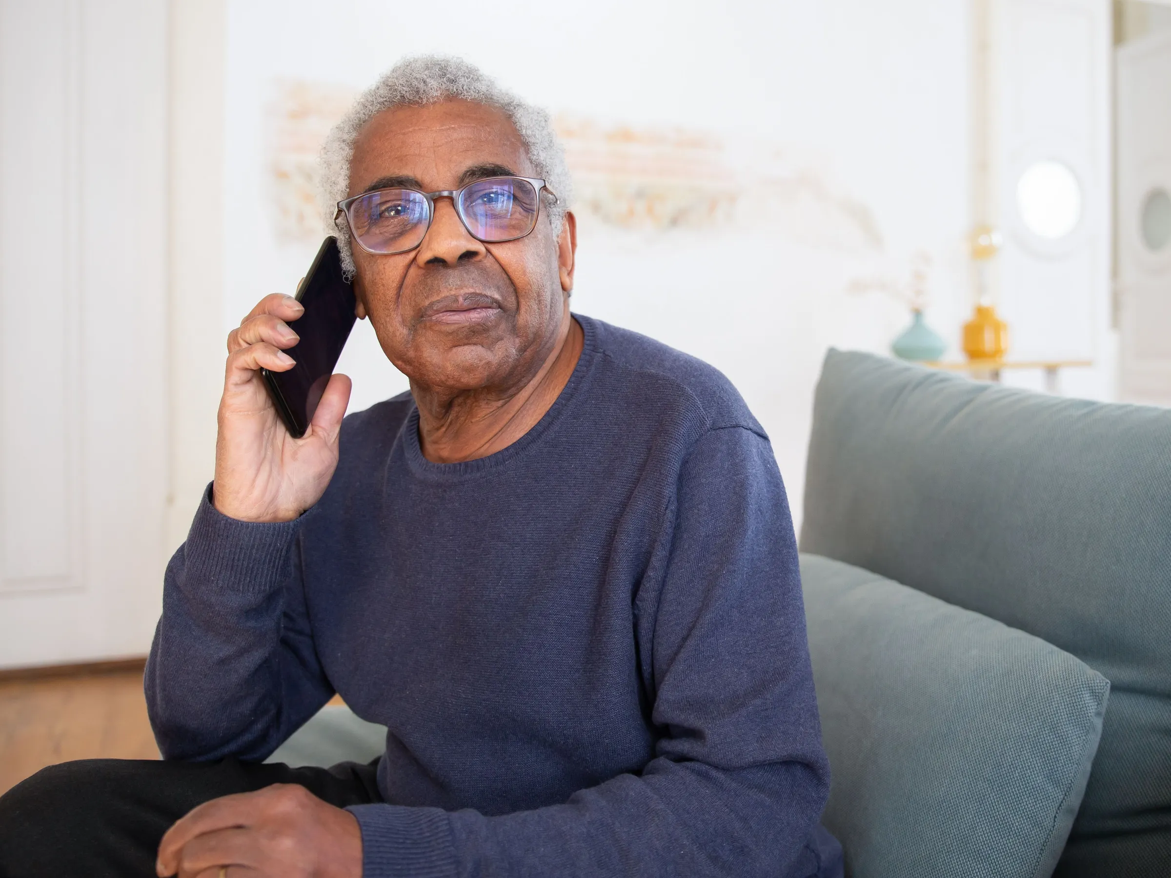An older man with gray hair and glasses sits on a sofa in a bright living room, talking on a mobile phone.