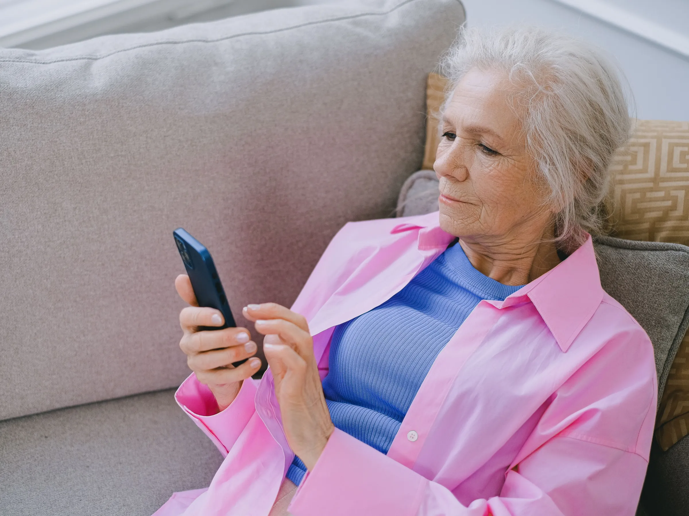 An older woman with white hair leans back on a gray sofa and looks at a smartphone.