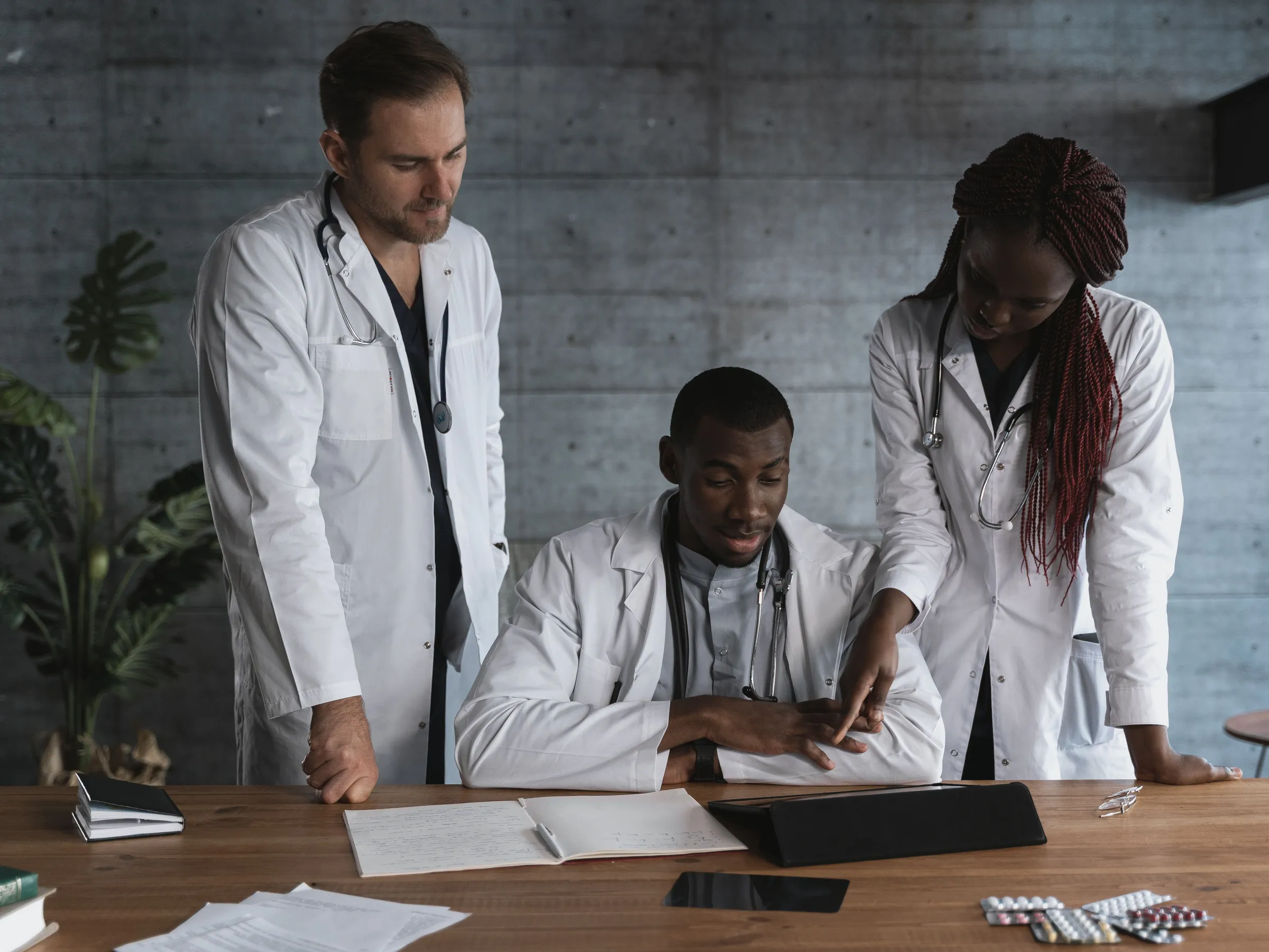 Three clinicians in white coats at a wooden table; one sits while two stand beside him, one pointing to a tablet.