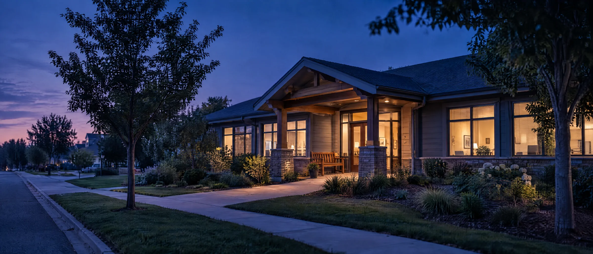 A small single-storey primary-care practice at blue hour, its windows glowing warm along a quiet, tree-lined street.